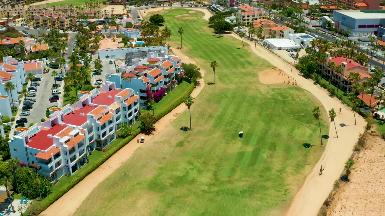 San Jose Del Cabo aerial frame with the fairway centered, a small golf cart lower in the course, red-roof buildings to the left, sandy path to the right, and no visible people.