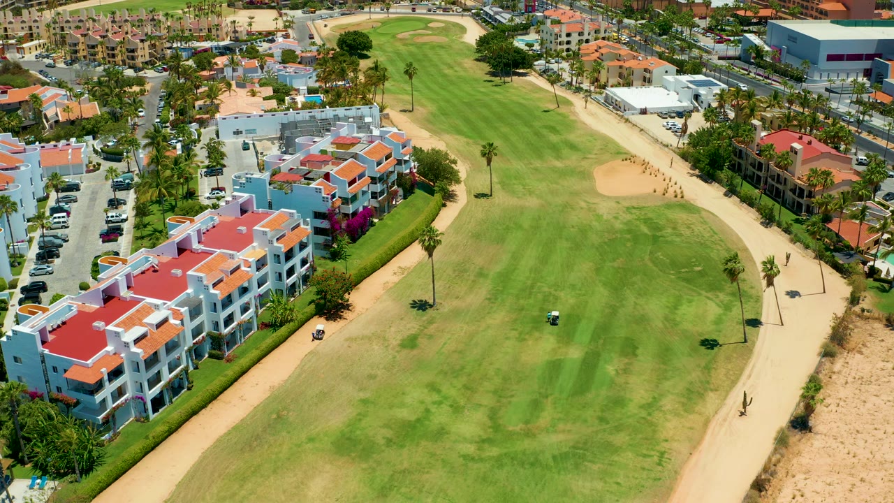 Aerial view in San Jose Del Cabo with a curved sandy path along the fairway, a small golf cart on the grass, red-roofed buildings on the left, palm trees, and no visible people.