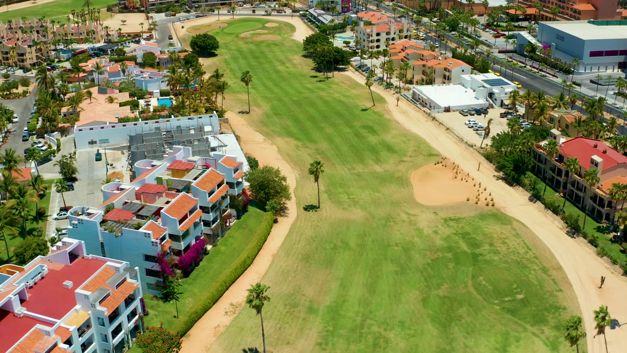 Aerial frame from San Jose del Cabo showing a green golf fairway, tan bunker, palm trees, white tent area, parked cars, and red-roofed residential buildings at the start of the clip.