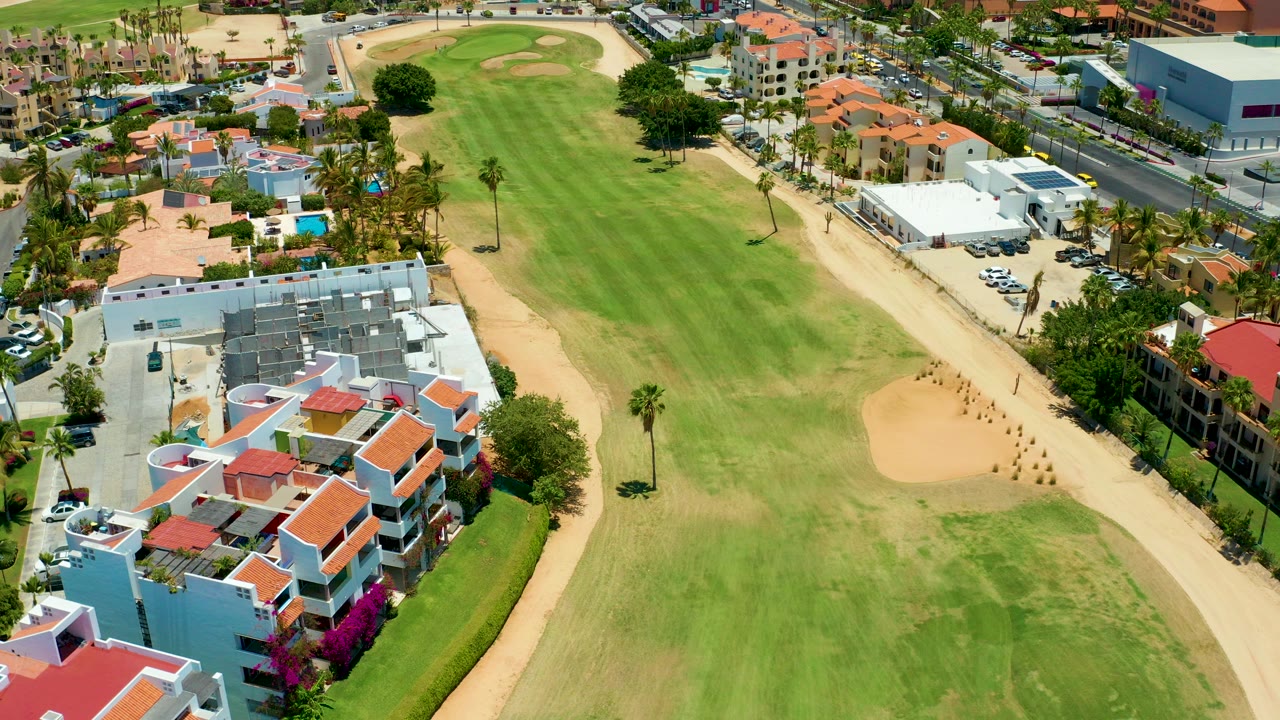High aerial frame over a San Jose del Cabo golf fairway with red-roofed homes in the lower left, tan bunkers, palm trees, surrounding streets, and no visible people.