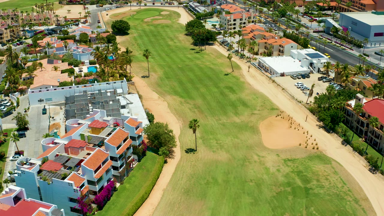 Drone frame over the San Jose del Cabo course with mowed green grass, tan sand areas, homes with red roofs, a roadside row of buildings, parked cars, and no visible people.