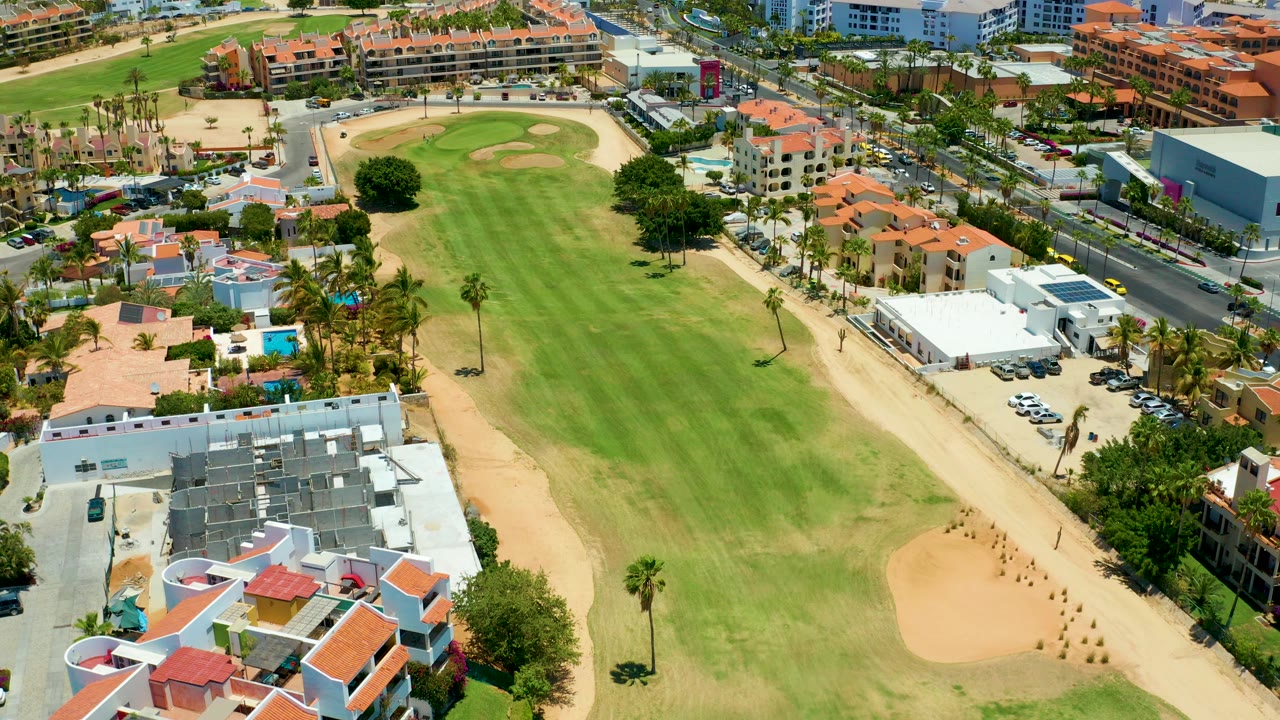 Aerial frame in San Jose del Cabo shows a long green golf fairway, tan sand bunkers, red-roofed buildings, palms, roads, parked cars, and no visible people.