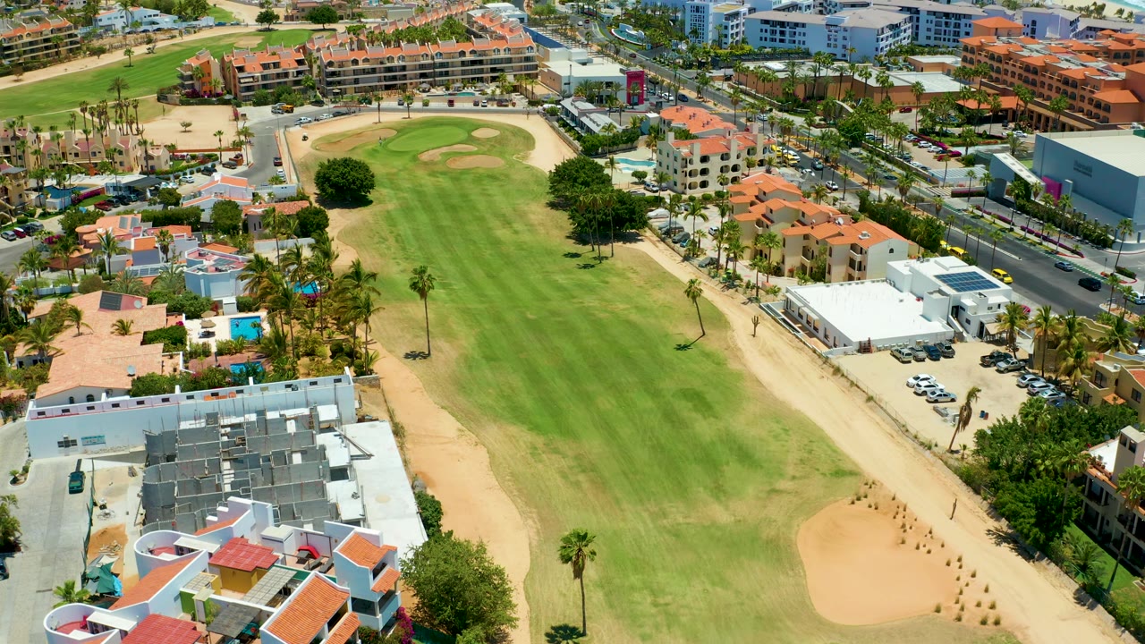 Aerial frame over San Jose del Cabo shows the golf course extending through the center, with palms, roadside cars, red-roofed buildings, white structures, sand areas, and no visible people.