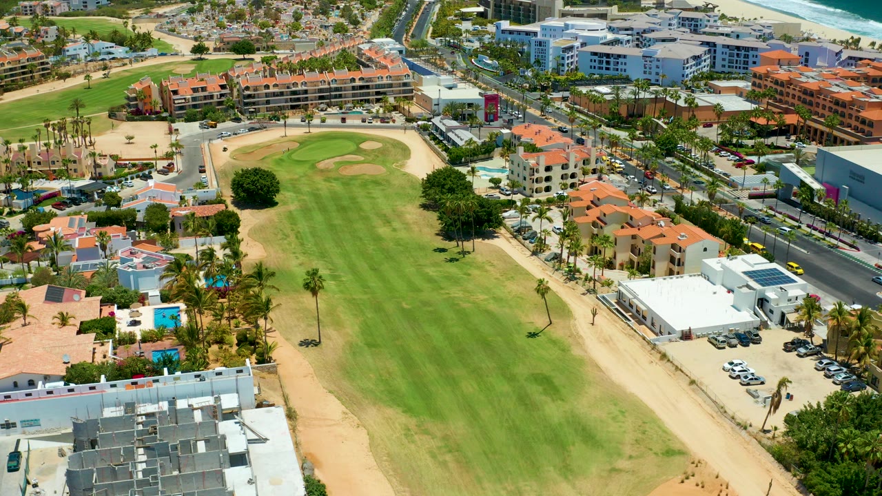 Aerial frame from San Jose del Cabo showing a broad green golf fairway bordered by sandy paths, palms, red-roof buildings, road traffic, and a distant ocean strip, with no visible people.