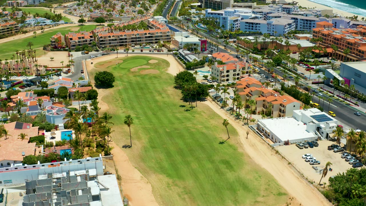 Aerial frame from San Jose del Cabo showing striped fairway grass, sandy course edges, palms, a roadside row of red-roof buildings, light traffic, and a bright ocean band, with no visible people.