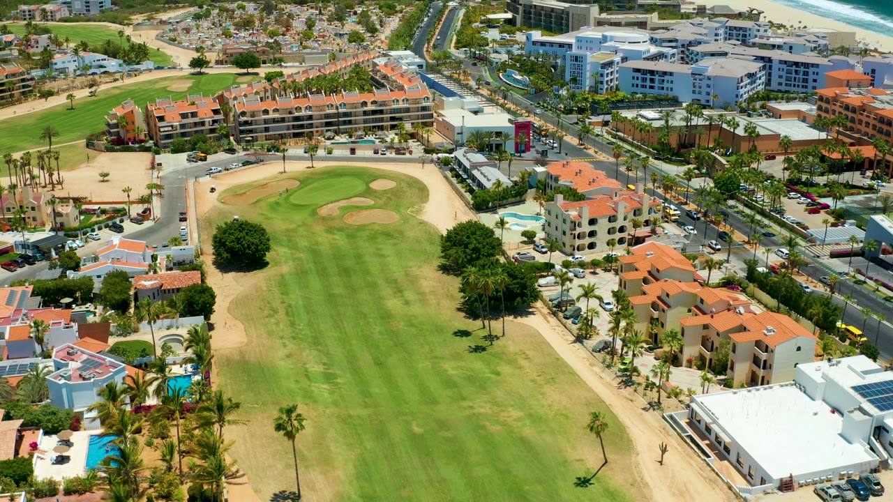 Opening aerial frame over San Jose Del Cabo showing a green fairway with sand traps, red-roofed resort buildings, road traffic, larger buildings, and the beach near the top edge, with no visible people.