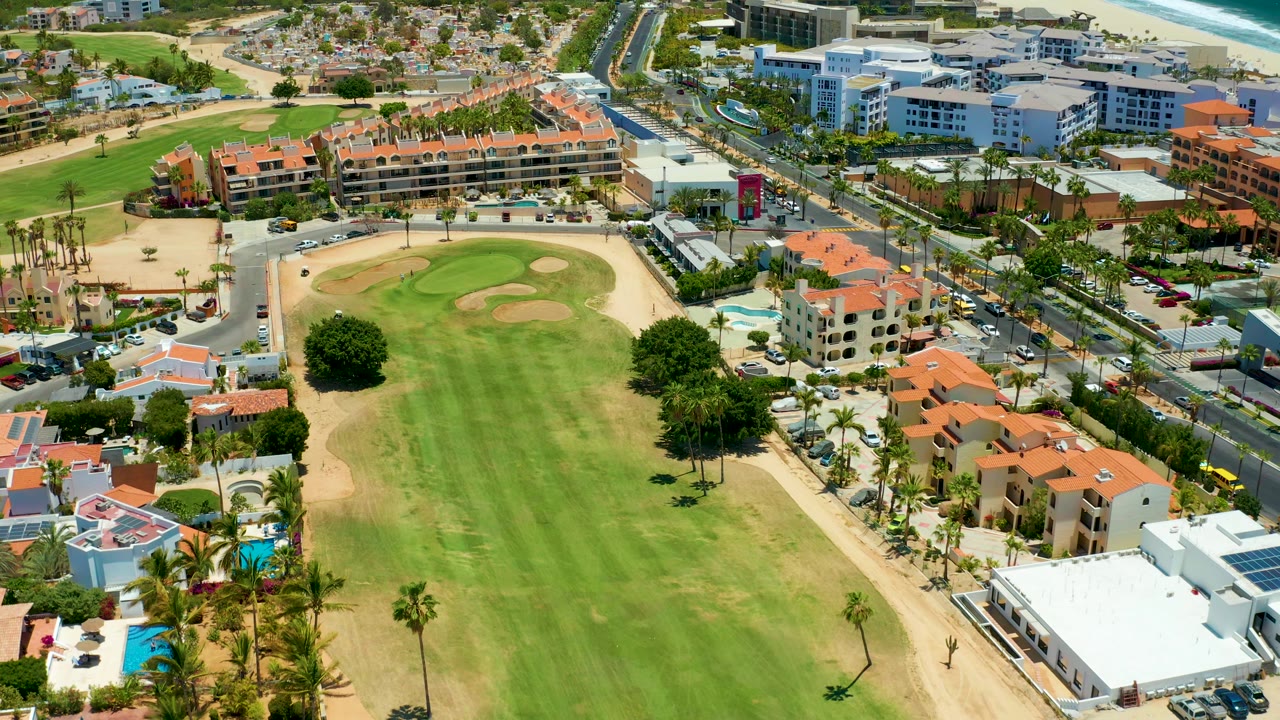 San Jose Del Cabo aerial frame with the fairway centered, tan bunkers at midframe, villas and pools in the lower right, cars on the road, and beachfront buildings in the distance, with no visible people.