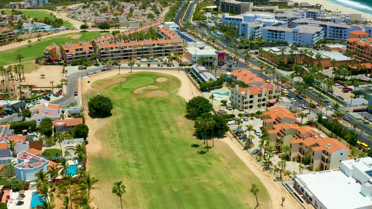 San Jose Del Cabo resort aerial showing a broad golf green in the foreground, a cluster of red-roofed buildings to the right, moving cars on the bordering road, and a narrow ocean strip beyond the development, with no visible people.