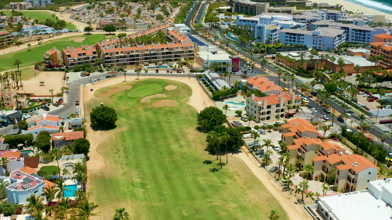 Drone frame looking over a golf course, road intersection, palm trees, and resort buildings.
