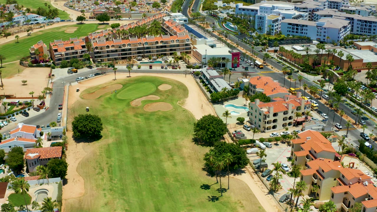 Aerial frame over San Jose del Cabo showing a green fairway, tan sand traps, bordering roads, red-tiled buildings, parked cars, and no visible people.