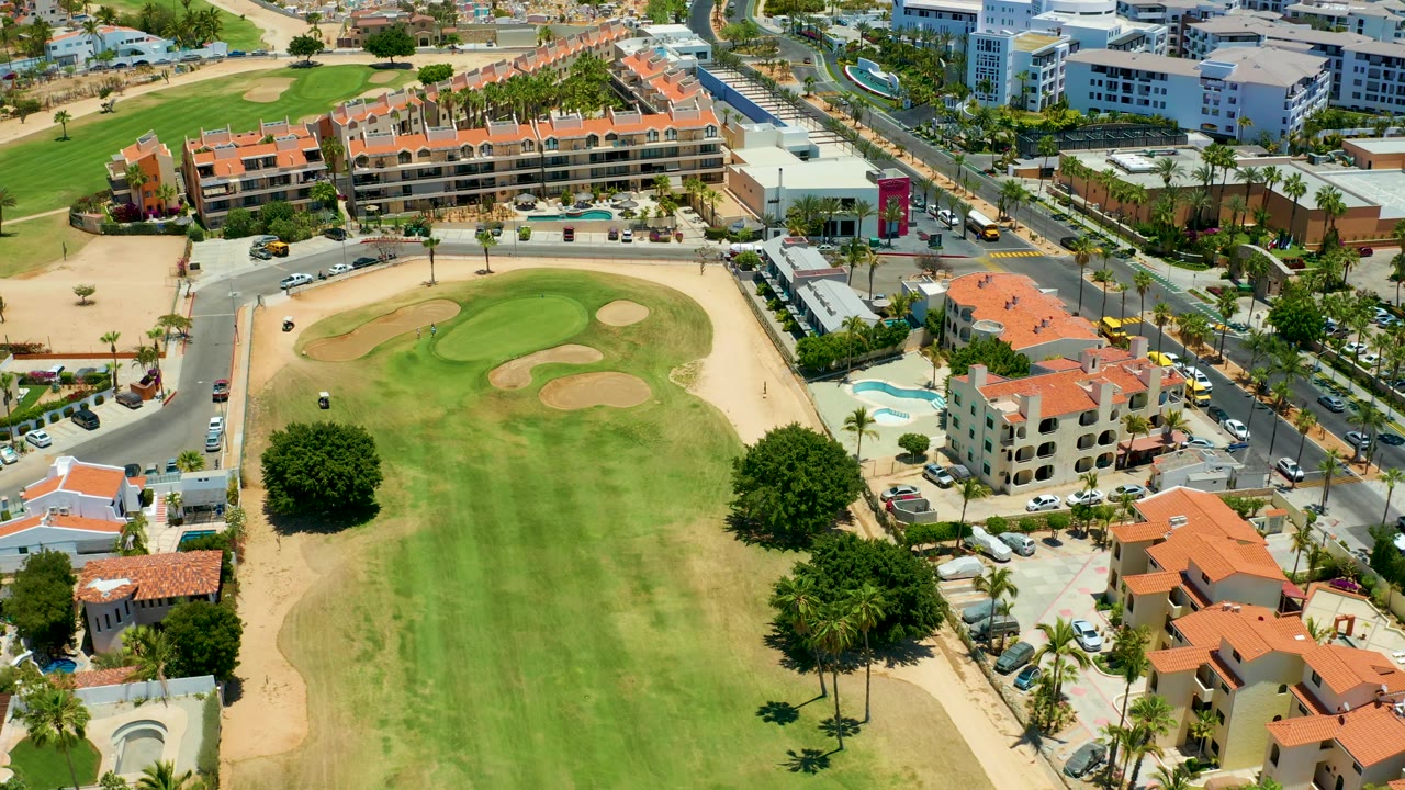 Elevated San Jose del Cabo frame showing a golf green and sand traps beside a curved road, parked vehicles, palm-lined properties, and no visible people.