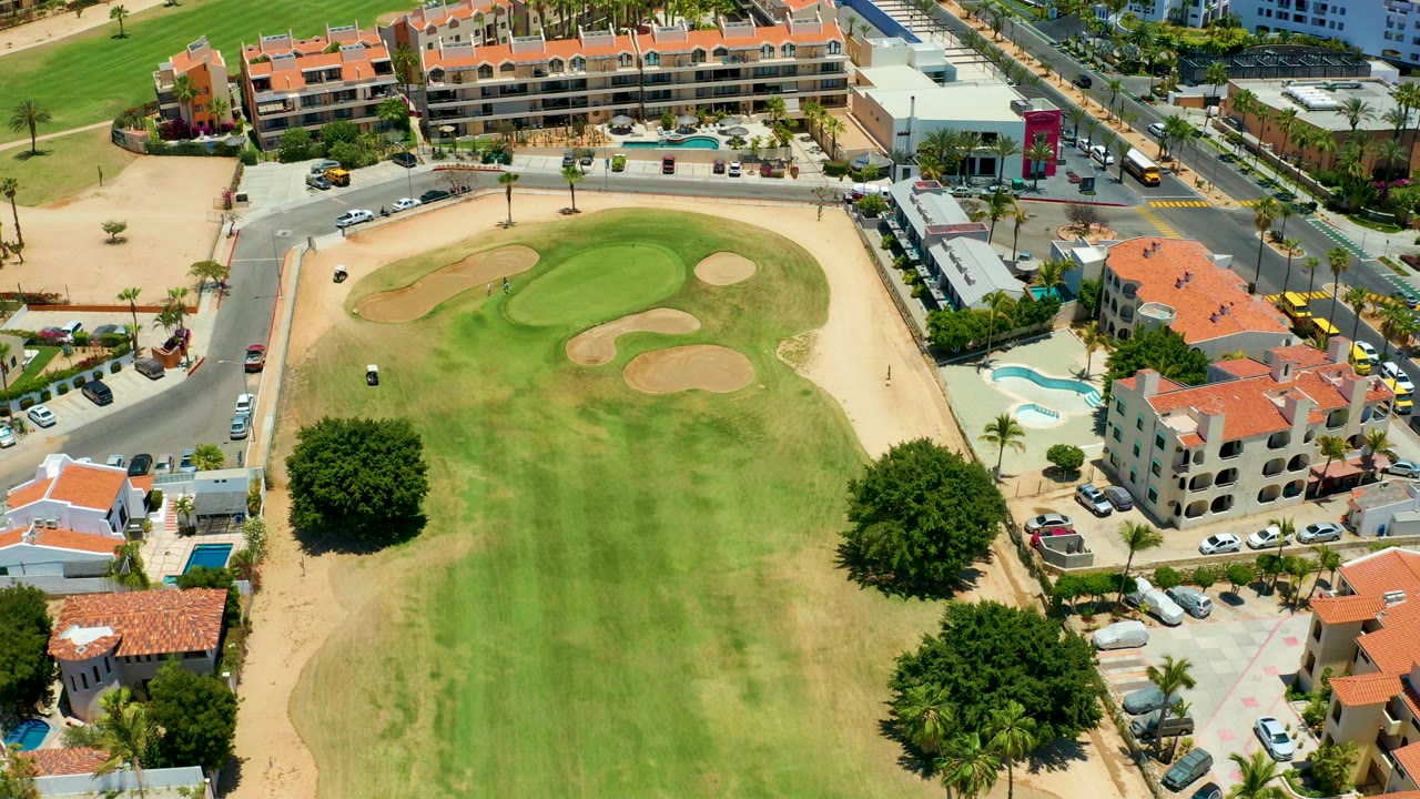 Opening aerial frame of a San Jose Del Cabo golf course with fairway grass, sand traps, a small golf cart, red-roof homes, cars, palms, and no visible people.