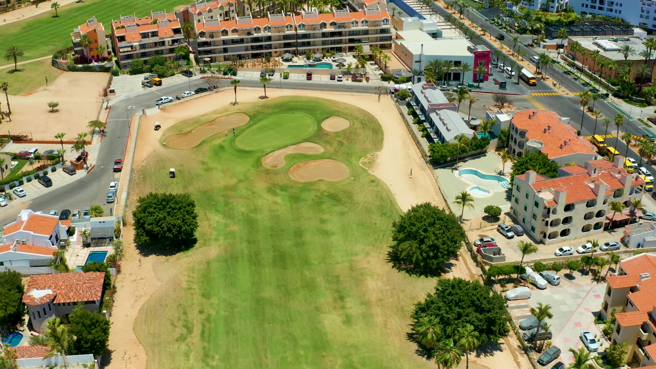 Aerial frame in San Jose Del Cabo showing the golf cart farther across the fairway, with sand traps near the top, red-roof buildings, cars on roads, and no visible people.
