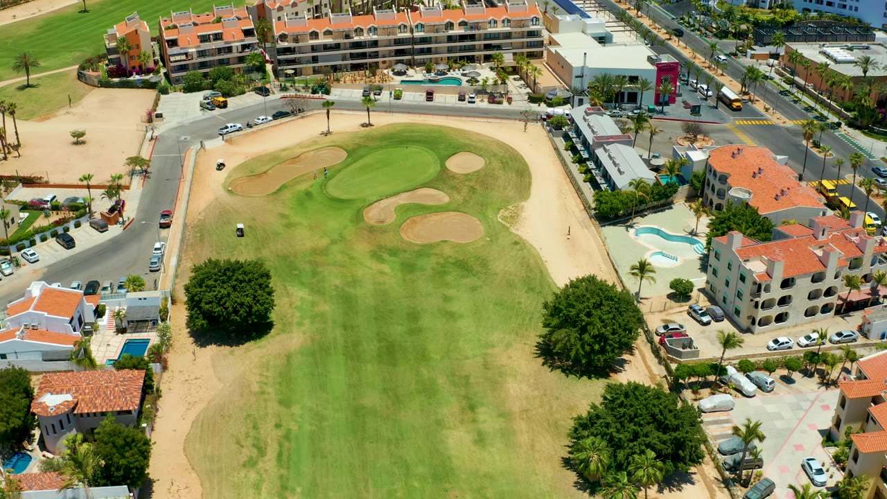 San Jose Del Cabo aerial frame with the cart near the left side of the course, green and tan fairway patches, sand traps, roads, red roofs, and no visible people.