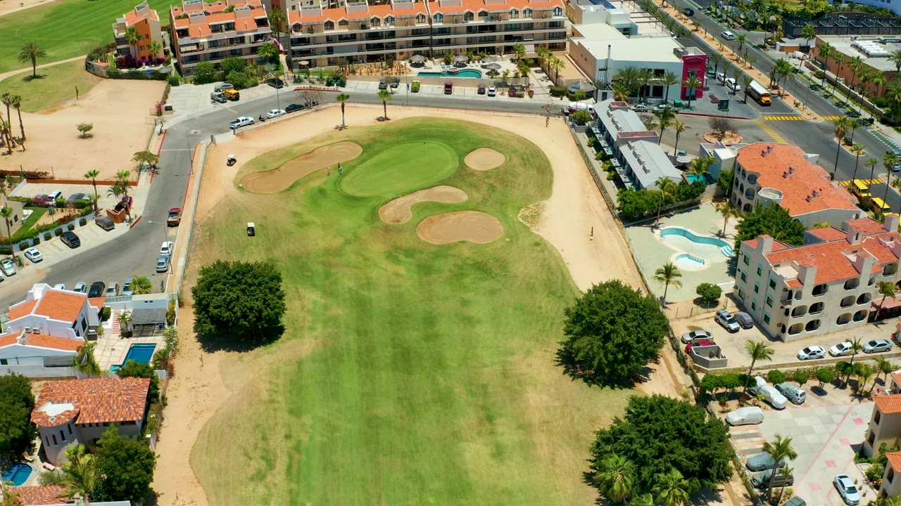 Drone view over a golf green bordered by streets, homes, cars, and a pool.
