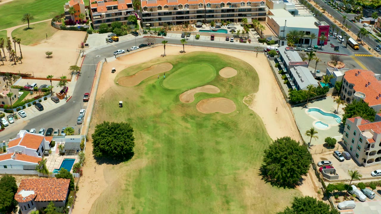 Overhead 4K frame of a San Jose del Cabo golf green with sand traps, dry rough, adjacent roads, parked cars, low-rise buildings, pools, and no visible people.