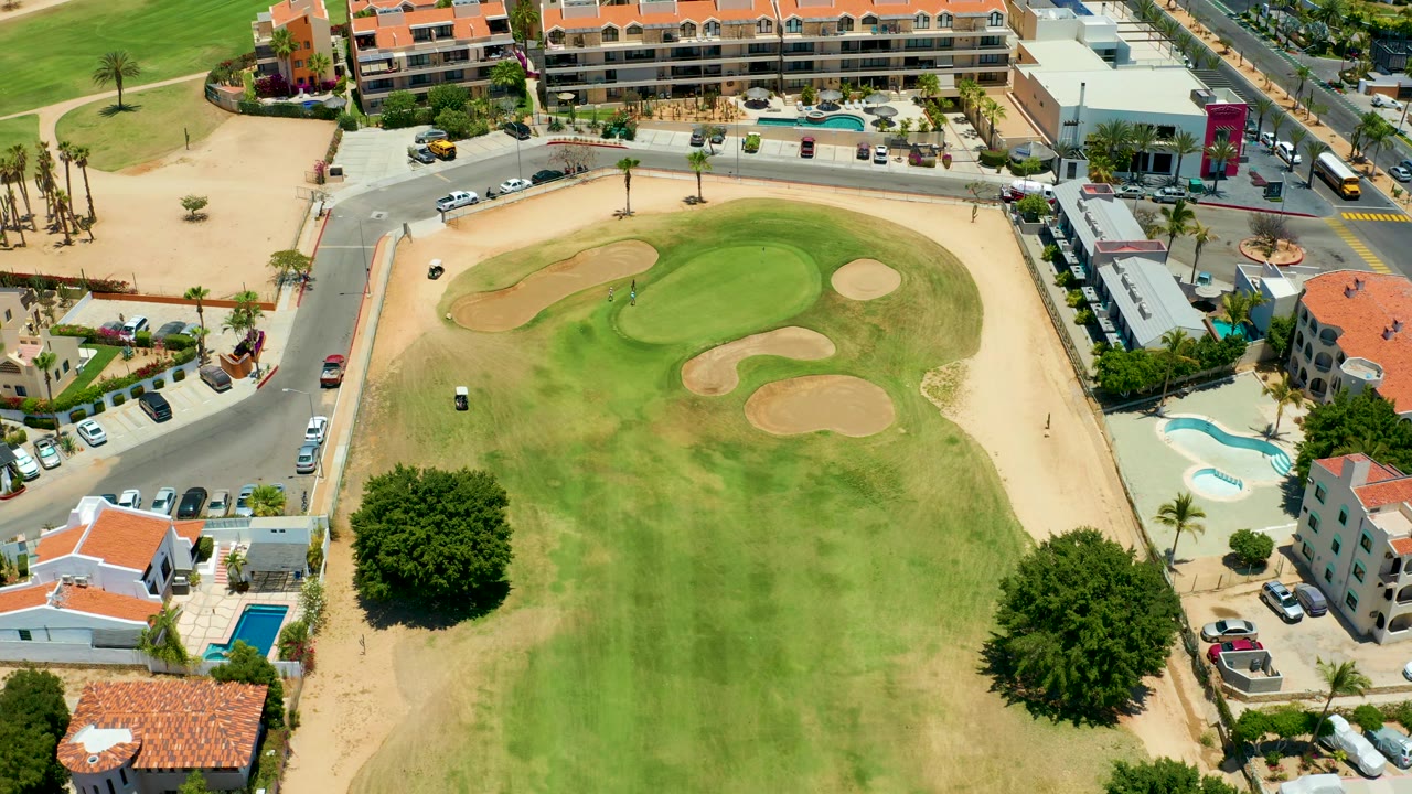 High-angle San Jose del Cabo frame with the golf cart lower on the green, several sand traps ahead, road traffic above the course, low-rise buildings, palms, pools, and no visible people.
