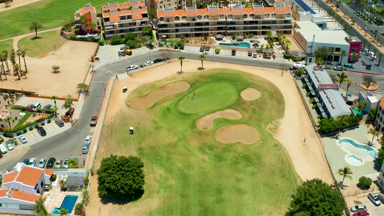 Aerial frame over a San Jose Del Cabo golf course with sand traps, a small golf cart on the green, roads, cars, palm trees, pools, and low buildings, with no visible people.
