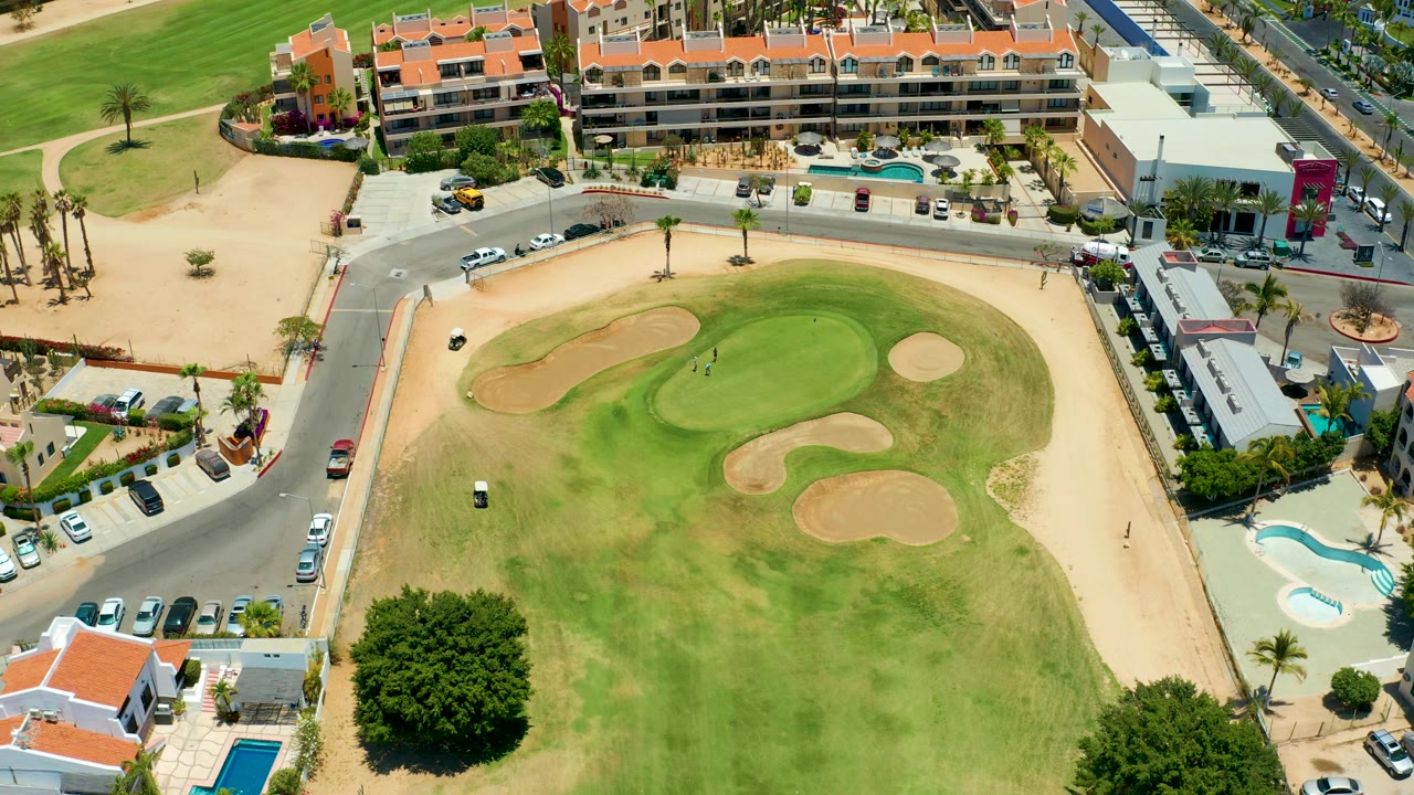 High oblique aerial frame of the San Jose Del Cabo course with the green centered, sand traps around it, a golf cart on the grass, roads and cars along the edges, and no visible people.