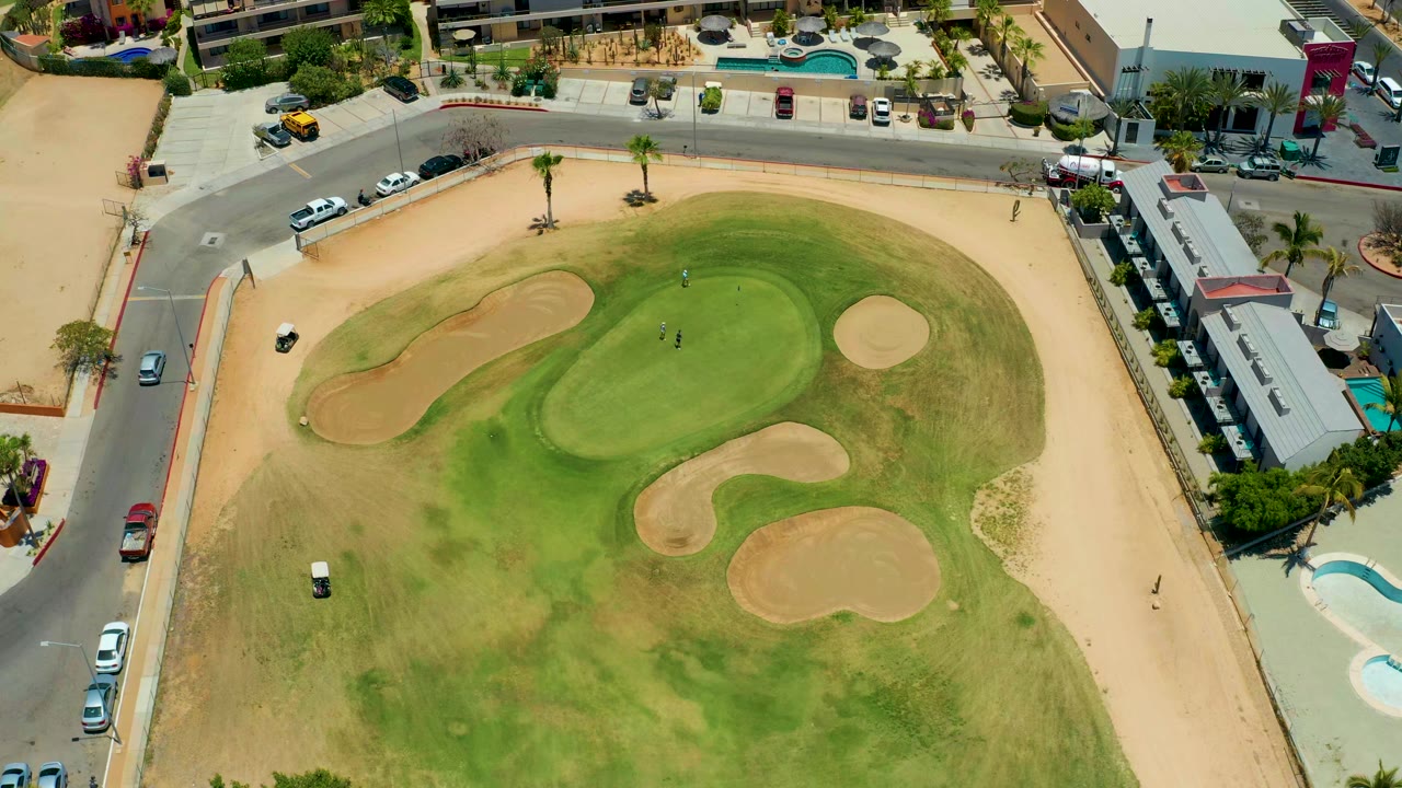 Aerial frame in San Jose del Cabo with the putting green centered, bunkers around it, a curved road across the top, vehicles near the curb, buildings at the edge, and no clearly visible people.