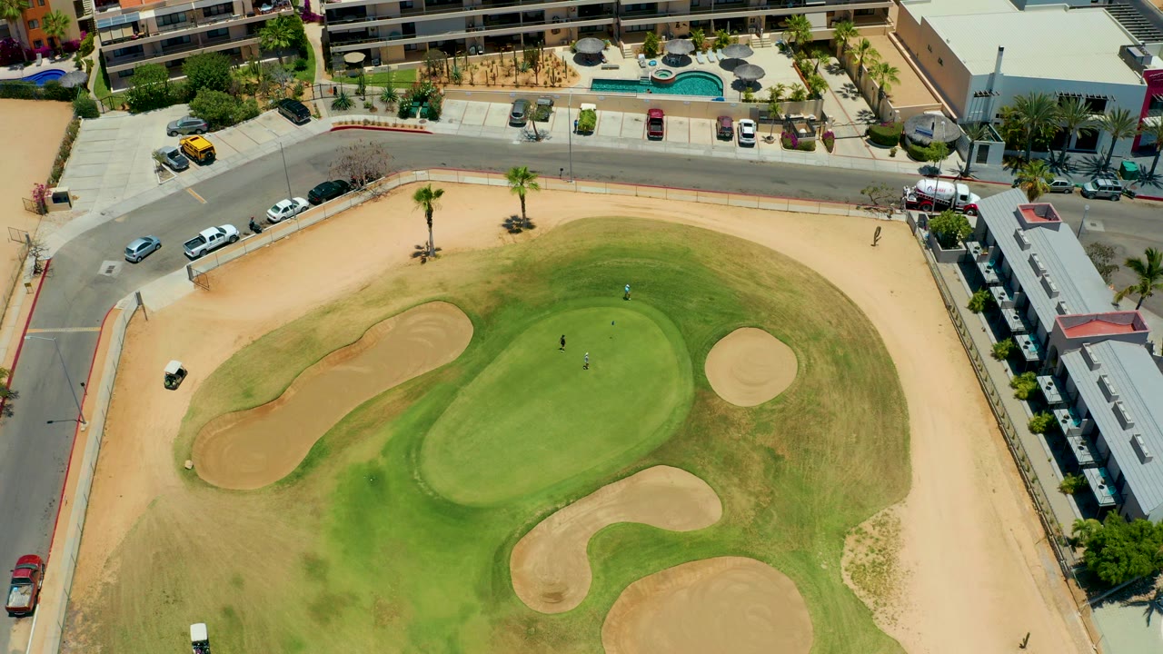 Overhead view in San Jose del Cabo of a golf green ringed by tan bunkers, dry rough, palm trees, roadside parking, and small visible people on the course.