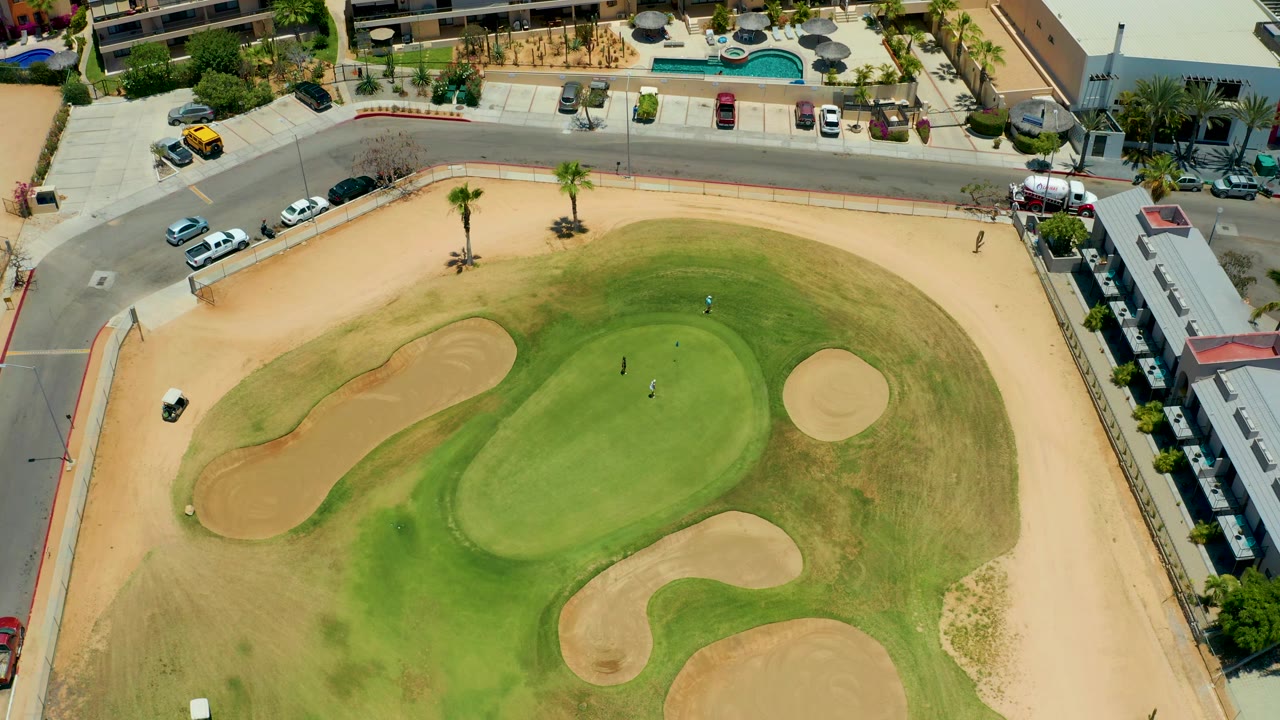 High overhead view in San Jose del Cabo with the green shifted across frame, tan bunkers around it, a sandy border, roadside cars, palms, and small people on the turf.