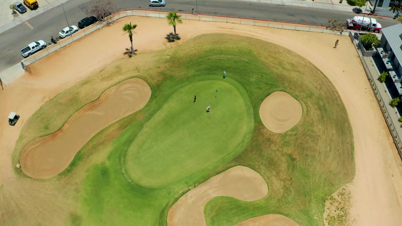 Aerial golf green with golfers, bunkers, cart path, palms, and roadside cars.