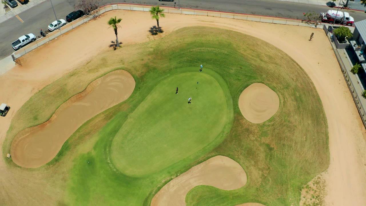Drone-style overhead view in San Jose Del Cabo with the putting green filling the center, a round bunker on the right, larger sand traps on the left and bottom, golfers on the grass, palms, and cars by the road.