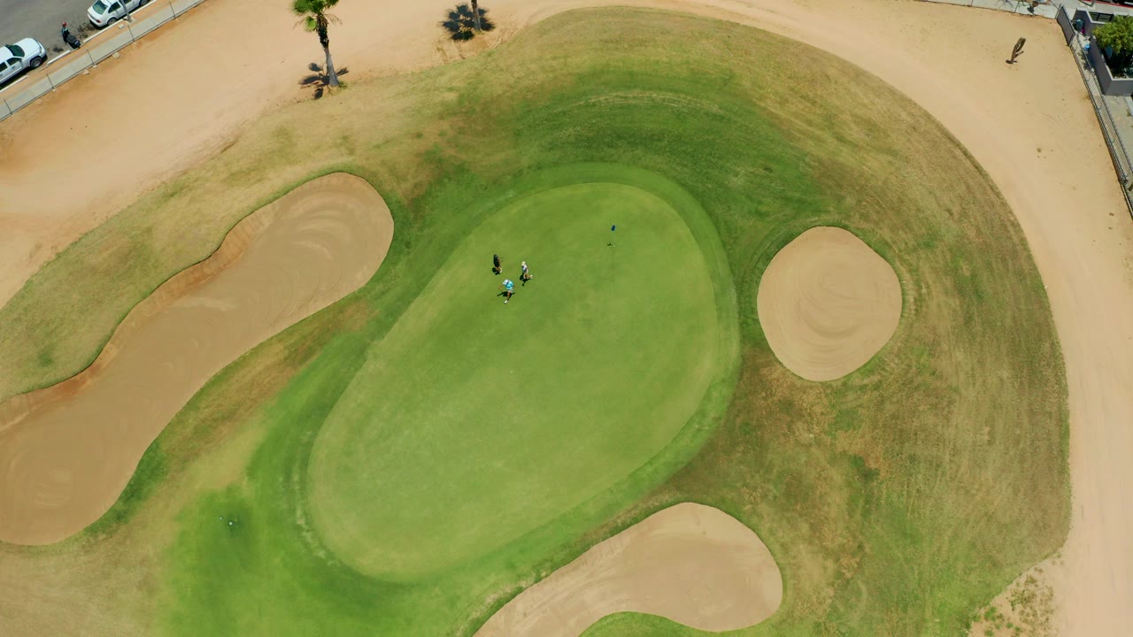 Overhead view in San Jose Del Cabo, Baja California Sur, showing a putting green encircled by sand bunkers and small golfers near the upper center of the green.