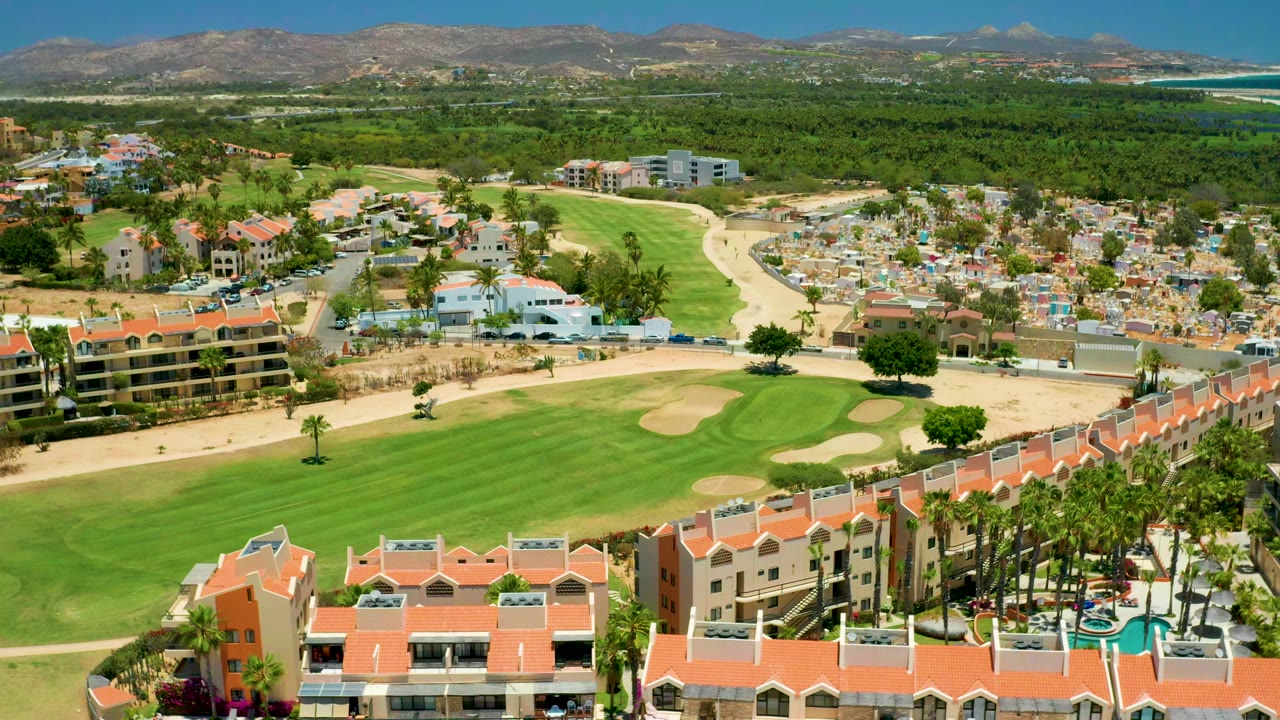 Aerial frame from San Jose del Cabo showing red-roofed residential buildings in front, green fairway and sand traps at left, a cemetery at right, distant mountains, a blue sky, and no visible people.