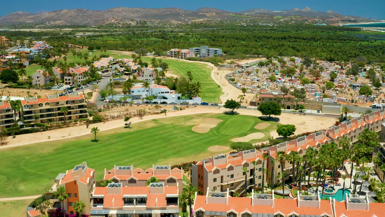 High aerial over golf fairway, sand traps, red-roofed buildings, cemetery, hills, and ocean