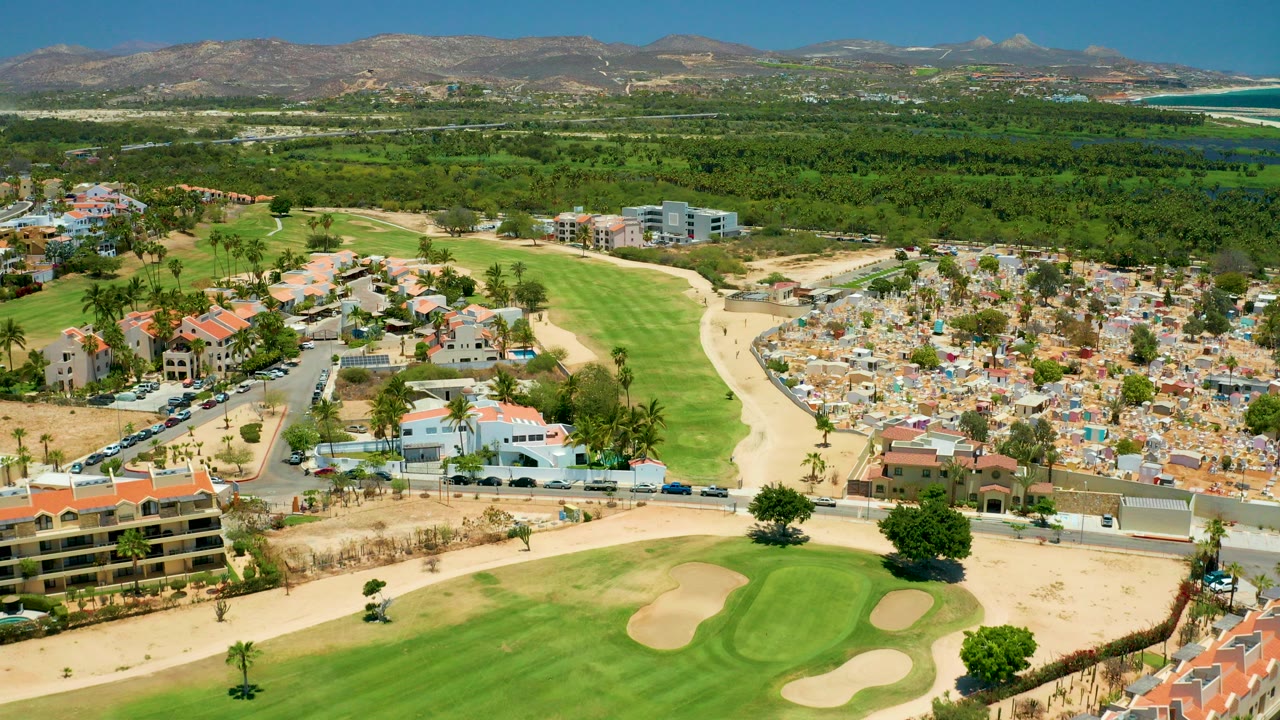 Aerial view over San Jose Del Cabo with golf fairways and sand traps in the foreground, a dense residential area to the right, ocean and mountains in the distance, and no visible people.
