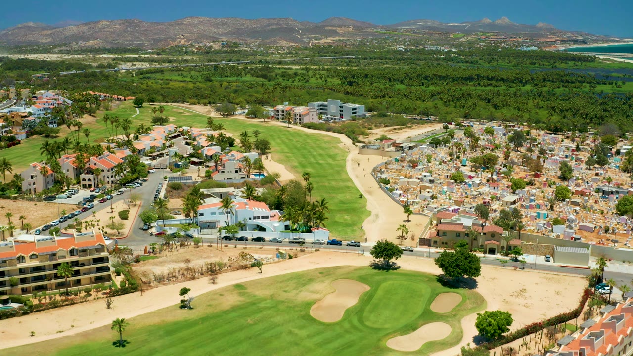 Drone view of green fairway beside residential blocks and distant coastline.
