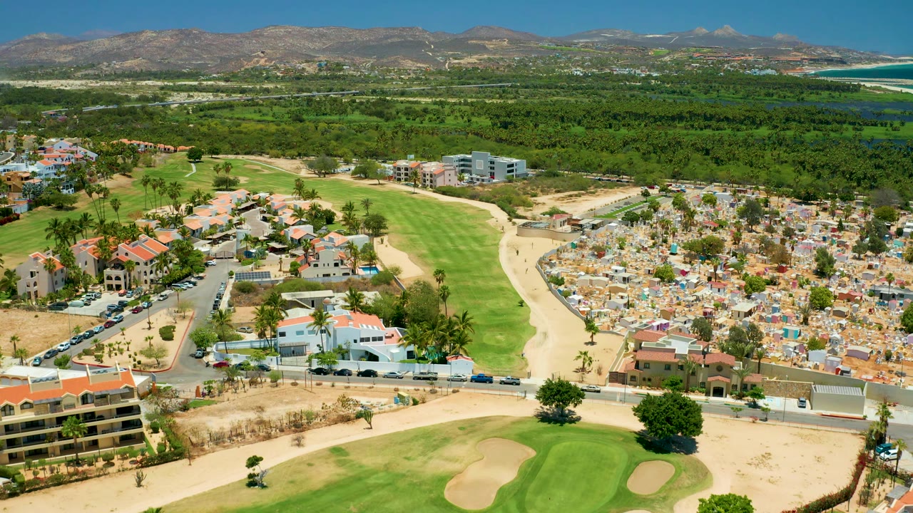 Wide San Jose Del Cabo aerial showing trimmed golf greens and bunkers in front, compact residential buildings to the side, beach and blue ocean beyond, distant mountains, and no visible people.