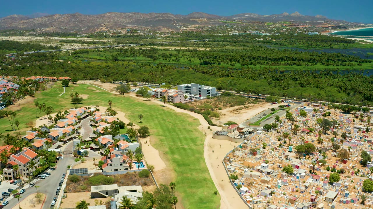 San Jose del Cabo aerial view with the fairway more centered, red-roof houses at lower left, cemetery-like grounds at lower right, green vegetation beyond, mountains on the horizon, and no visible people.