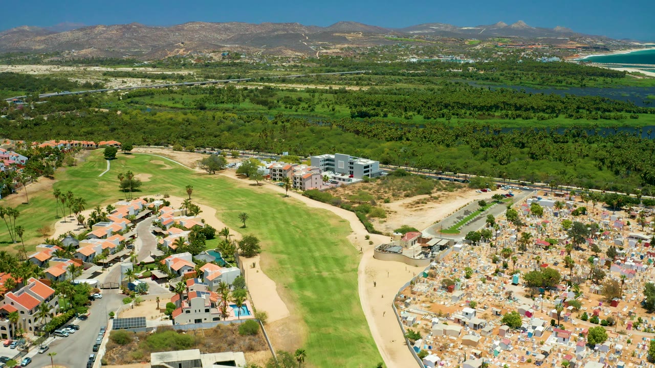 A wide San Jose del Cabo drone frame shows golf greens, tan sand paths, clustered homes, cemetery-like grounds, a broad line of trees, mountains, and a strip of blue coastal water, with no visible people.