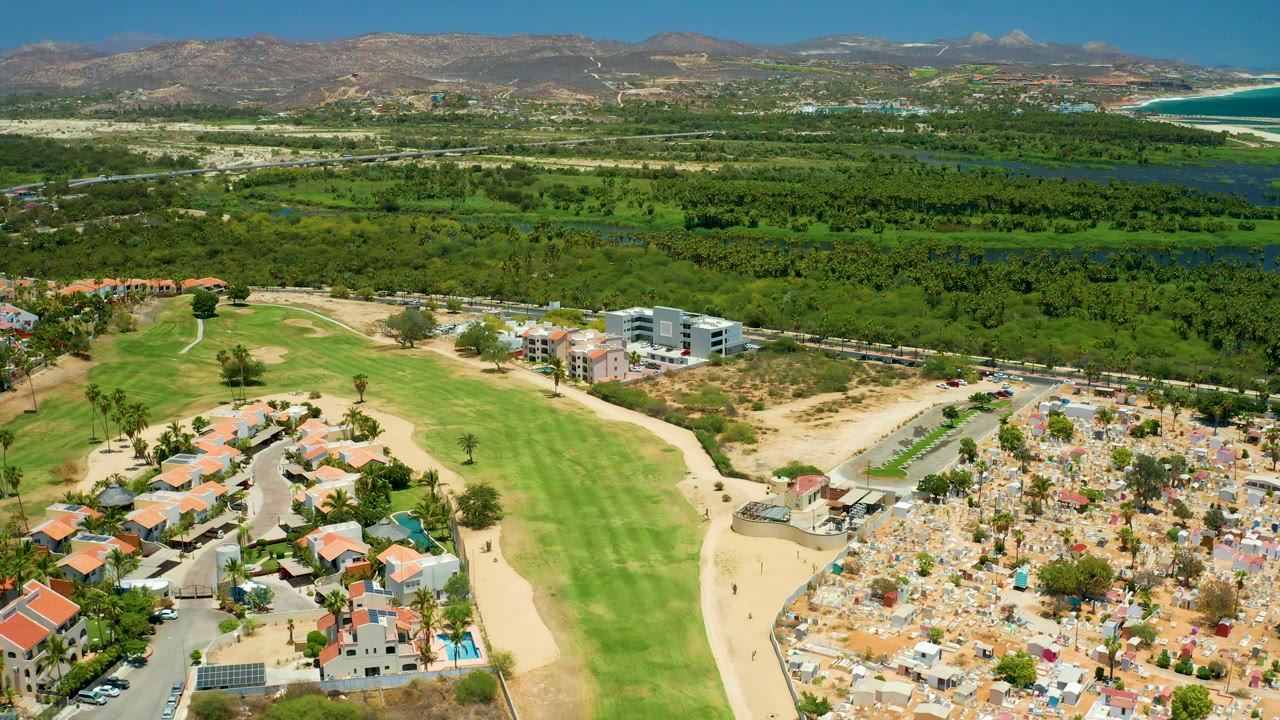 Aerial pan frame with fairway, homes, cemetery grounds, modern buildings, dense trees, mountains, and water.