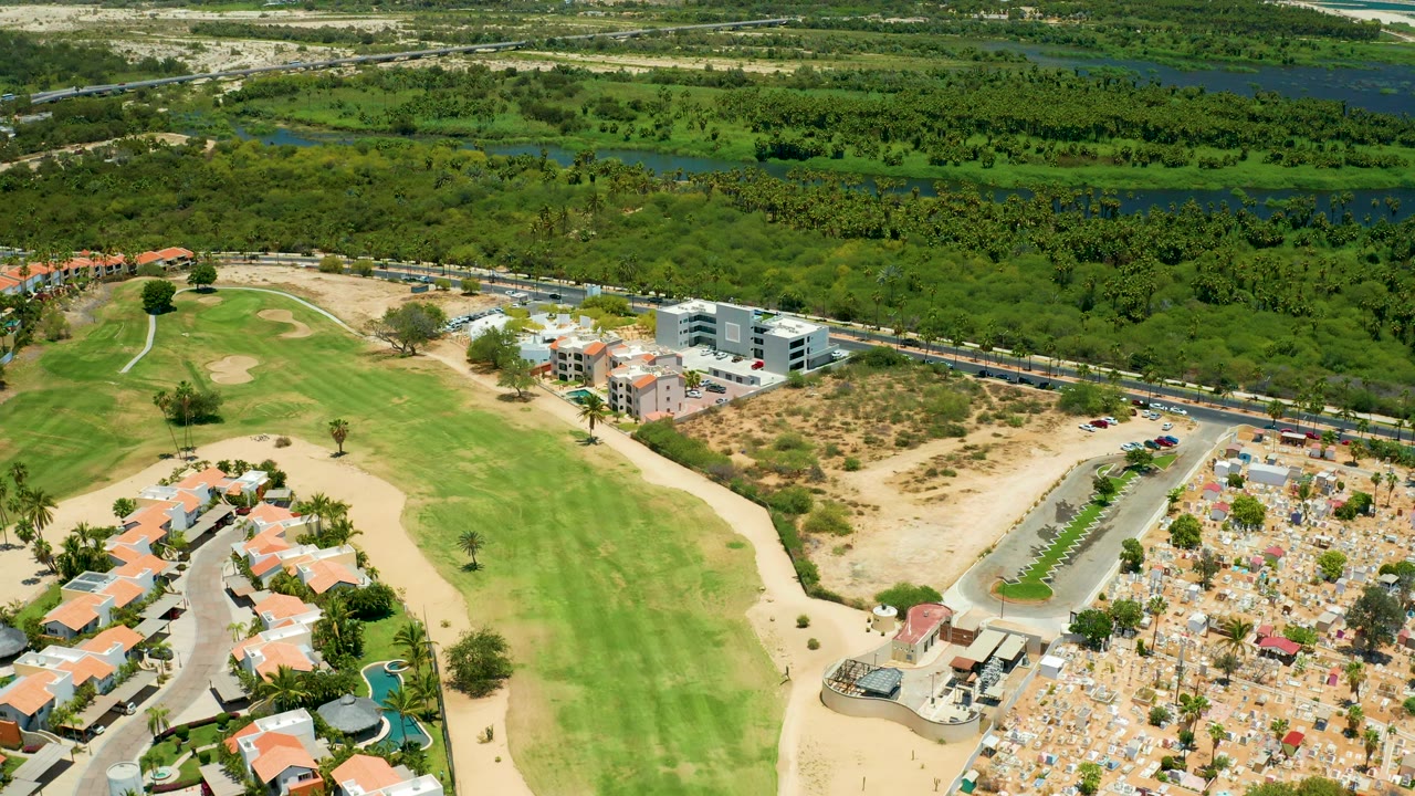 San Jose del Cabo frame with the golf course filling the lower center, red-tiled homes on the left, a modern light-colored building near the road, and a green waterway corridor across the top, with no visible people.