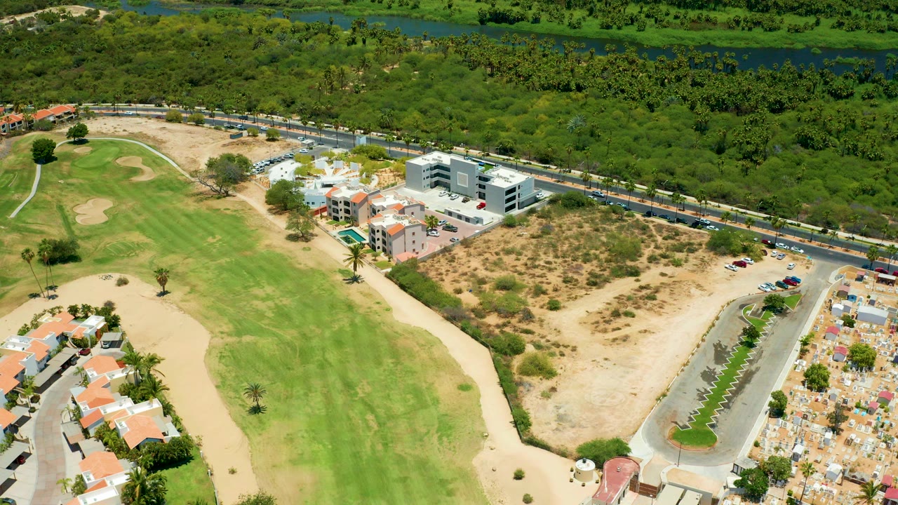 Opening aerial frame in San Jose Del Cabo shows a green golf fairway, sand traps, a dry lot, nearby homes, a road with vehicles, dense trees, and no visible people.