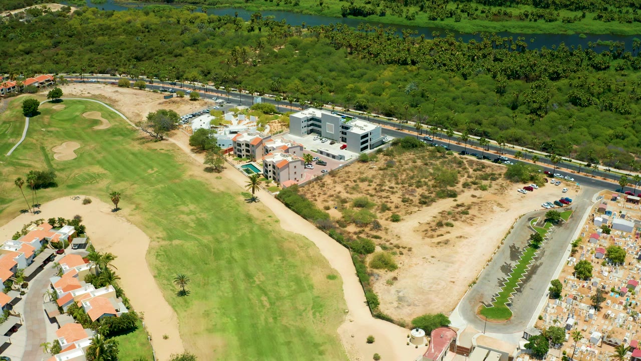 At 0:01 the San Jose Del Cabo aerial view centers the fairway and dry lot while the road, cars, modern buildings, homes, trees, and no visible people remain in frame.