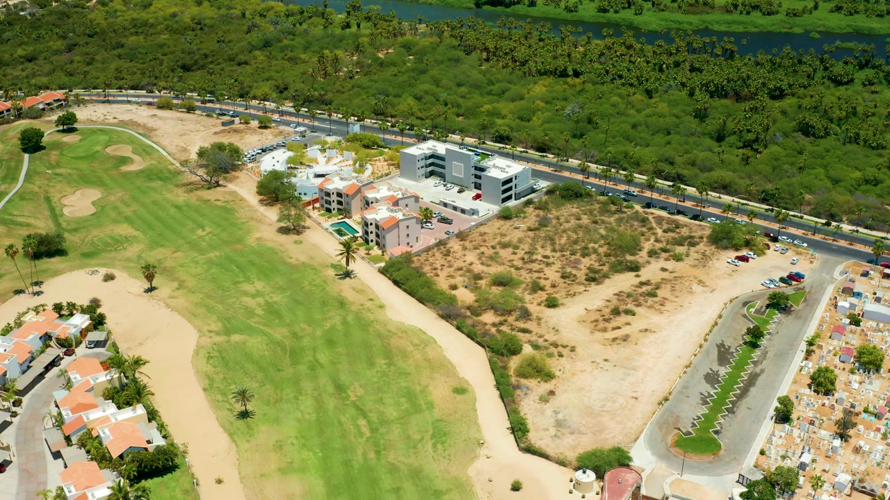At 0:02 the San Jose Del Cabo frame shows the fairway running diagonally past sand traps, a roadside line of vehicles, residential buildings, a parking area, thick greenery, and no visible people.