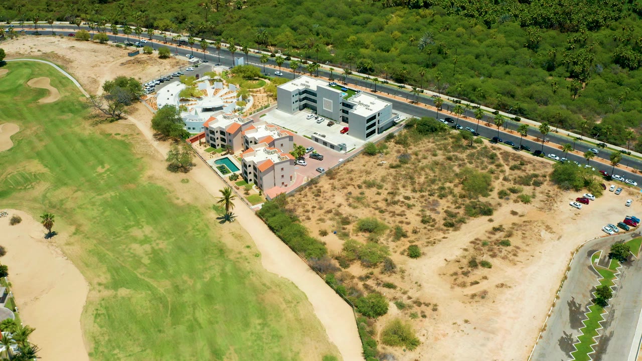 Aerial view in San Jose Del Cabo showing a green golf fairway, dry scrub land, resort buildings, a pool, road traffic, and dense trees with no visible people.