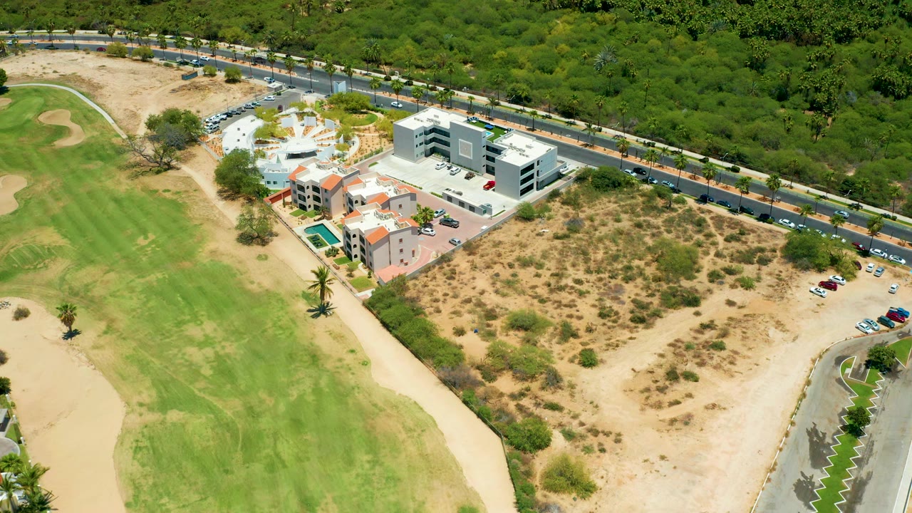 Elevated view of cars on a road beside golf turf, resort buildings, pool, and scrub land.