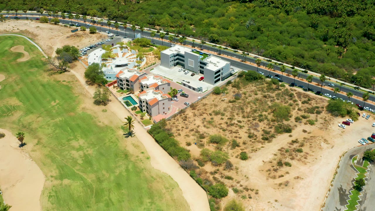 At 0:02, the aerial view in San Jose Del Cabo keeps the road traffic at the upper edge, with resort buildings, a pool, dry land, golf grass, and no visible people.