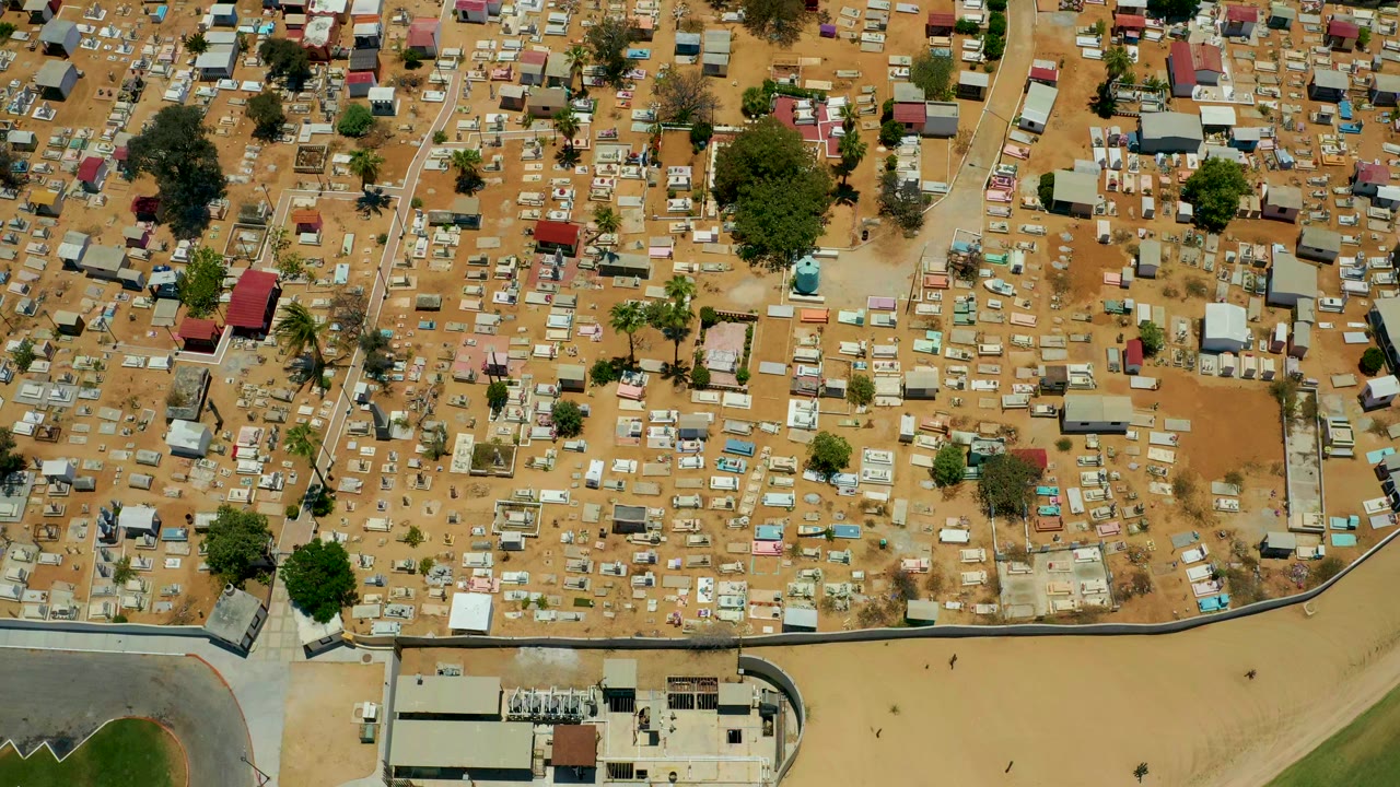 Overhead frame from San Jose Del Cabo, Baja California Sur, showing dense small structures and rectangular plots on sandy ground near a walled edge, with no visible people.
