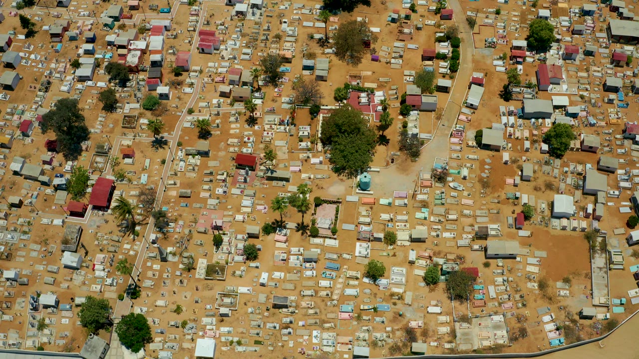 Overhead frame of San Jose del Cabo showing dense small huts, pale roofs, sandy dirt lanes, scattered trees, and no visible people at the start of the clip.