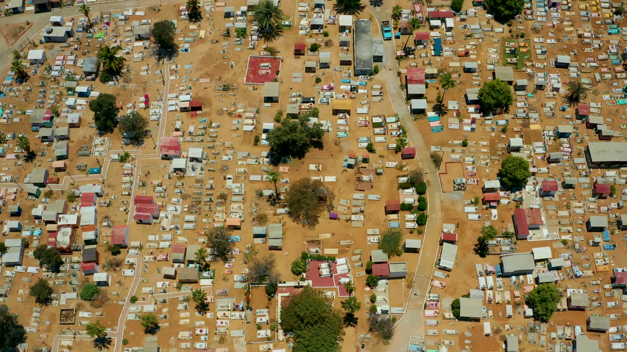 High overhead view in San Jose Del Cabo, Baja California Sur, showing dense small huts and structures on tan sandy ground with scattered trees, dirt paths, and no visible people.