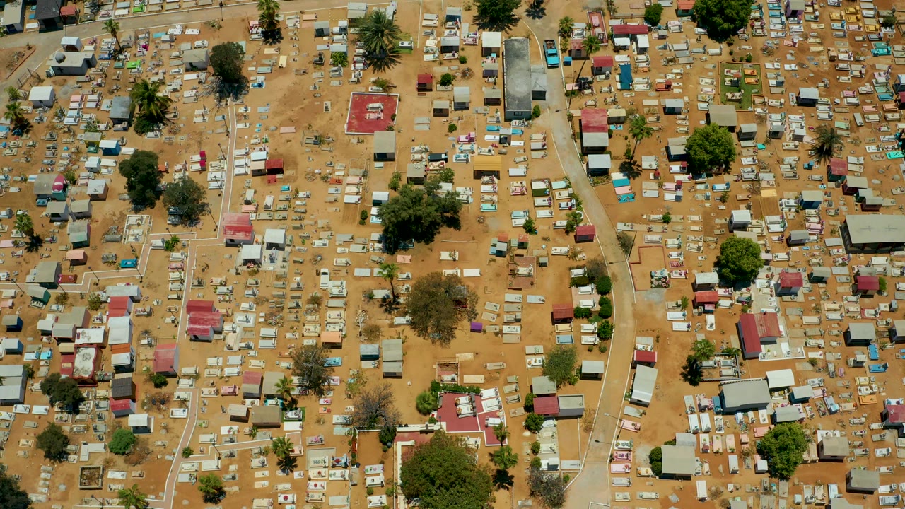 Top-down frame over San Jose Del Cabo with many small roofs and hut-like structures arranged across sandy terrain, broken by narrow dirt lanes and no visible people.