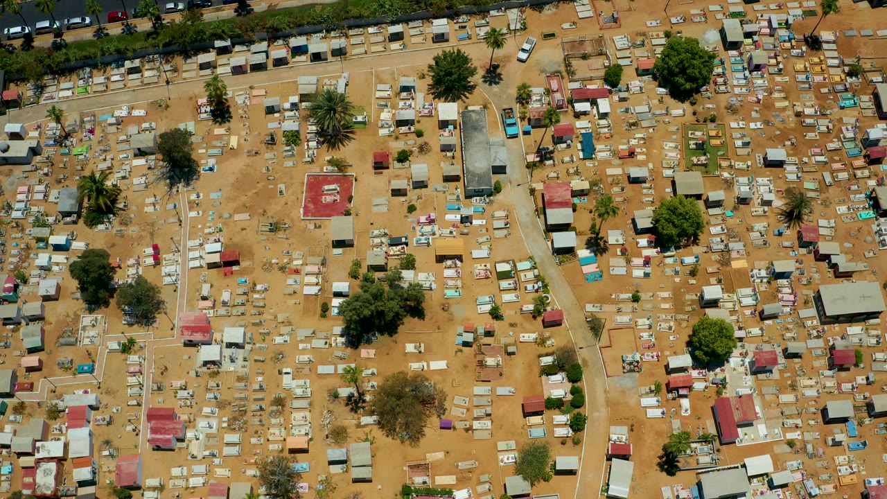 High overhead frame of a dry informal settlement in San Jose del Cabo, Baja California Sur, with many small huts, sandy ground, trees, paths, and no visible people.
