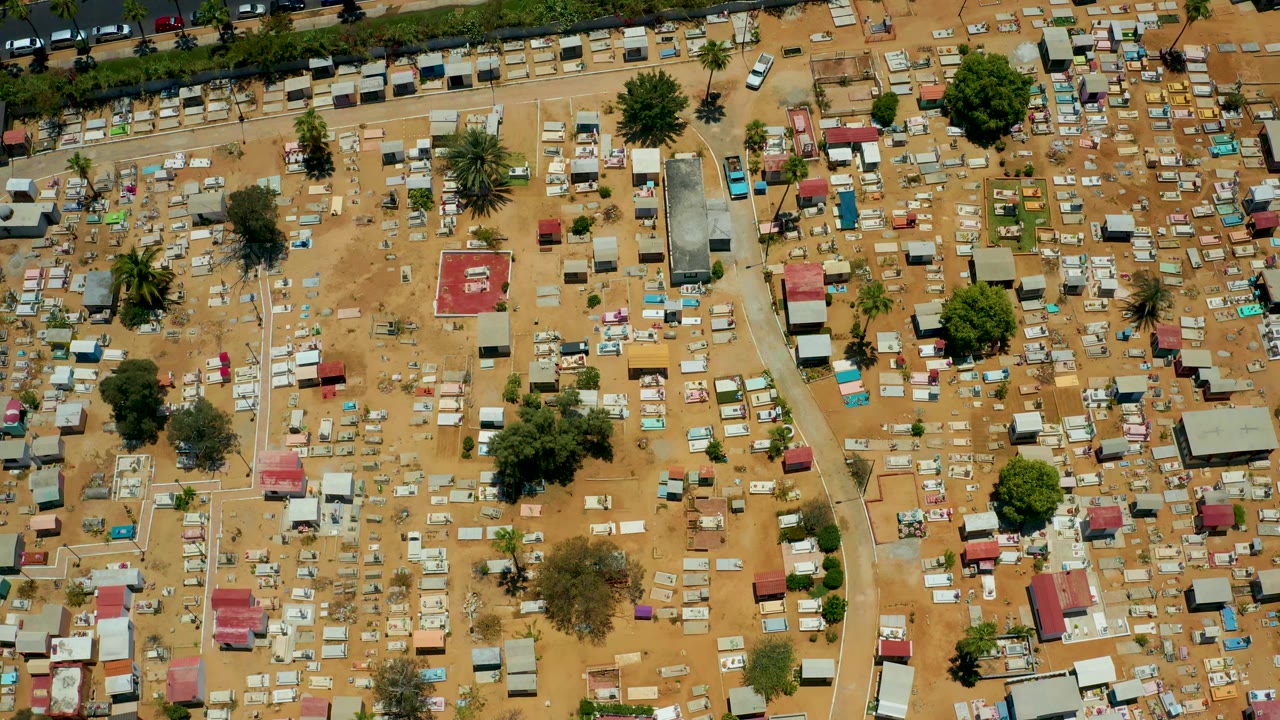 Overhead aerial frame in San Jose del Cabo showing clustered huts, dusty footpaths, a tree-lined road edge at the top, and no visible people.